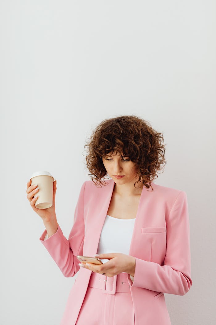 Confident woman in a pink blazer holding coffee and texting indoors.